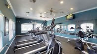 This fitness center is thoughtfully designed to support a well-rounded workout routine. The space is outfitted with a row of cardio machines, including treadmills and ellipticals, positioned to take advantage of natural light streaming in through a series of arched windows along the walls. The windows not only brighten the interior but also create an open, airy atmosphere that enhances the exercise experience.

A floor-to-ceiling mirror spans a full wall, allowing users to monitor their form while also visually expanding the space. Adjacent to the cardio area, a strength training station provides additional equipment for resistance-based workouts. The flooring is a durable, dark rubberized material well-suited for high-impact activity.

The ceiling features recessed lighting and dual ceiling fans, ensuring consistent air circulation and a comfortable environment throughout the room. A wall-mounted flat-screen television adds an entertainment element for those looking to stay engaged during longer workout sessions.

The walls are painted in a bold teal tone, lending the space an energetic and motivating ambiance. Crown molding along the ceiling adds a polished architectural detail, elevating the overall finish of the fitness center beyond purely functional design.