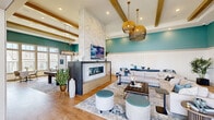 This generously proportioned single-floor community space offers an open-concept layout designed for both relaxation and social gathering. The main lounge area commands attention with soaring ceilings accented by exposed wood beams and bold teal upper walls that transition to crisp white wainscoting below. A floor-to-ceiling marble fireplace serves as the dramatic focal point of the room, paired with a wall-mounted TV and flanked by stylish seating arrangements on warm wood-look flooring. Woven rattan pendant lights add a coastal, organic touch throughout the space.

The full kitchen features light wood cabinetry, a large quartz-topped island with bar seating, stainless steel appliances, open shelving, and matching pendant lighting—ideal for catering and community events. A bright dining nook with floor-to-ceiling windows floods the space with natural light and offers views of the surrounding outdoor amenities.

A covered outdoor patio connects seamlessly to the main lounge through wide sliding doors, equipped with its own TV and comfortable lounge chairs, creating a smooth indoor-outdoor flow. Additional seating nooks and a billiards area round out the amenities. Large windows throughout the space invite abundant natural light and frame views of lush exterior landscaping, including palm trees.