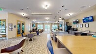 Located at 400 Katrina Ct in Midlothian, VA, this community clubroom at 288 Lofts is a thoughtfully designed amenity space spanning a generous single floor. The open-concept layout is anchored by a large granite-topped kitchen island with round upholstered bar stools, complemented by dark wood cabinetry, a full-size refrigerator, and a dedicated countertop service area — ideal for community gatherings and events. Stylish pendant lighting in a mix of matte black and globe styles hangs overhead, adding a contemporary flair to the space. The flooring transitions between light wood-look planks near the kitchen and patterned area rugs throughout the lounge zones, creating distinct yet cohesive areas. Multiple seating arrangements feature mid-century modern-inspired sofas, accent chairs, and small side tables in warm caramel and neutral tones. Wall-mounted flat-screen televisions are positioned throughout the room for easy viewing from multiple vantage points. Large windows and glass double doors along the perimeter flood the interior with natural light and provide views of the lush greenery and outdoor pool area. The warm, cream-toned walls and carefully curated artwork contribute to an inviting, upscale ambiance throughout.
