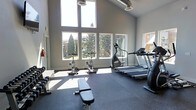 This fitness center is a thoughtfully designed, single-floor workout facility that combines functionality with an open, airy atmosphere. The space is defined by its impressively high ceilings and large, stacked windows that flood the interior with abundant natural light, offering pleasant views of surrounding trees and greenery — creating an inviting environment for exercise.

The layout is well-organized and spacious, with durable rubber flooring throughout that provides both safety and comfort during workouts. Cardio equipment is prominently featured, including multiple treadmills and an elliptical trainer positioned to take advantage of the natural light and outdoor views. A dedicated cycling area houses stationary spin bikes, ideal for high-intensity interval or endurance training sessions.

A strength training section offers a weight bench and a dumbbell rack, providing the essentials for resistance and free-weight workouts. Exposed ductwork along the upper ceiling adds an industrial design element that complements the clean, modern grey-toned walls. The overall ambiance is bright, clean, and motivating — a well-rounded facility that caters to a variety of fitness routines in a comfortable and visually appealing setting.