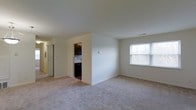 This single-floor residence offers a clean and functional layout designed for comfortable everyday living. The main living area is spacious and open, featuring soft carpeted flooring in a neutral tone that extends throughout the space, creating a warm and cohesive feel. The room is filled with natural light courtesy of a large window that looks out toward a lush, tree-lined exterior, bringing a sense of openness and connection to the outdoors. Smooth, white walls and a uniform ceiling height give the space a simple, uncluttered aesthetic that is easy to personalize. Adjacent to the living area, the kitchen is tucked neatly into its own defined space, showcasing dark espresso-toned cabinetry paired with a tiled backsplash that adds a touch of contrast and visual interest. The dark appliances complement the cabinetry for a cohesive look. A hallway connects the main areas of the residence, providing an efficient and intuitive flow throughout the floor plan. Recessed vents and subtle architectural details reflect thoughtful construction. Overall, the interior presents a move-in-ready environment with a neutral palette that serves as an ideal canvas for any décor style.