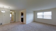 This single-floor residence offers a clean and functional layout designed for comfortable everyday living. The main living area is spacious and open, featuring soft carpeted flooring in a neutral tone that extends throughout the space, creating a warm and cohesive feel. The room is filled with natural light courtesy of a large window that looks out toward a lush, tree-lined exterior, bringing a sense of openness and connection to the outdoors. Smooth, white walls and a uniform ceiling height give the space a simple, uncluttered aesthetic that is easy to personalize. Adjacent to the living area, the kitchen is tucked neatly into its own defined space, showcasing dark espresso-toned cabinetry paired with a tiled backsplash that adds a touch of contrast and visual interest. The dark appliances complement the cabinetry for a cohesive look. A hallway connects the main areas of the residence, providing an efficient and intuitive flow throughout the floor plan. Recessed vents and subtle architectural details reflect thoughtful construction. Overall, the interior presents a move-in-ready environment with a neutral palette that serves as an ideal canvas for any décor style.