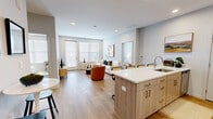 This thoughtfully designed two-story residence offers 2 bedrooms and 2 bathrooms across a spacious and functional floor plan. The main floor is expansive, housing the primary living areas, while the upper floor provides a more intimate setting for additional living or sleeping quarters.

The open-concept kitchen and living area anchors the main floor. The kitchen features light wood cabinetry, stainless steel appliances — including a refrigerator, range, and microwave — and a large island with a white quartz countertop and bar seating for multiple guests. Light-toned hardwood flooring runs throughout, complementing the soft gray walls and recessed lighting overhead.

The adjacent living room is bright and welcoming, with large windows fitted with horizontal blinds that allow abundant natural light to fill the space. A glass-paned door leads to a private balcony offering outdoor views of the surrounding streetscape.

Both bedrooms are well-sized and serene, featuring large windows for natural light, soft neutral tones, and dedicated closets. The primary bedroom is a comfortable retreat with ample room for furnishings.

The two bathrooms are finished in a clean, modern style, each featuring white countertops, undermount sinks, and full mirrors. The overall aesthetic throughout the residence is contemporary and cohesive, with a neutral palette that creates a calm, livable atmosphere.