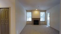 This single-floor residence offers a welcoming and well-appointed living space designed with both comfort and functionality in mind. The main living area serves as the focal point of the home, centered around a classic tile-surround fireplace with a clean mantel, providing a warm and inviting atmosphere. Soft, wall-to-wall carpeting in a neutral tone runs throughout, adding a sense of warmth underfoot and complementing the light-colored walls. Multiple windows flank the fireplace, flooding the room with abundant natural light and offering pleasant views of surrounding greenery. The recessed lighting above the fireplace niche adds a subtle architectural accent, highlighting the feature wall. The overall color palette is neutral and understated, with warm beige tones on the walls and ceilings creating a bright, airy ambiance. The open layout of the living area allows for versatile furniture arrangements, making it adaptable to a variety of living styles. A doorway visible to one side suggests additional rooms beyond, indicating a practical and well-organized floor plan. The space presents a clean, move-in ready condition with thoughtful details throughout.
