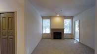 This single-floor residence offers a welcoming and well-appointed living space designed with both comfort and functionality in mind. The main living area serves as the focal point of the home, centered around a classic tile-surround fireplace with a clean mantel, providing a warm and inviting atmosphere. Soft, wall-to-wall carpeting in a neutral tone runs throughout, adding a sense of warmth underfoot and complementing the light-colored walls. Multiple windows flank the fireplace, flooding the room with abundant natural light and offering pleasant views of surrounding greenery. The recessed lighting above the fireplace niche adds a subtle architectural accent, highlighting the feature wall. The overall color palette is neutral and understated, with warm beige tones on the walls and ceilings creating a bright, airy ambiance. The open layout of the living area allows for versatile furniture arrangements, making it adaptable to a variety of living styles. A doorway visible to one side suggests additional rooms beyond, indicating a practical and well-organized floor plan. The space presents a clean, move-in ready condition with thoughtful details throughout.