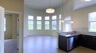 This single-floor space is defined by its standout architectural feature: a sweeping curved wall lined with a series of large windows that flood the interior with natural light and offer views of lush greenery outside. The soaring ceiling height amplifies the sense of openness, creating an airy, expansive atmosphere throughout the main living area. Light wood-toned flooring runs continuously across the space, complementing the neutral wall tones and contributing to a clean, contemporary aesthetic.

Pendant lighting and a centrally positioned drum-style ceiling fixture provide layered illumination, enhancing both the functional and decorative appeal of the space. The open layout allows the living area and kitchen to flow seamlessly together. The kitchen features dark cabinetry with a contrasting countertop, a stainless steel sink, and a stainless steel dishwasher, offering a sleek, modern finish. Additional pendant lights hang above the kitchen area, reinforcing the cohesive design language.

Natural light is a defining characteristic throughout, with windows positioned at varying heights to maximize brightness. The overall ambiance is modern, bright, and welcoming, with thoughtful architectural details that elevate the everyday living experience.