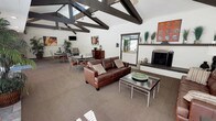 This single-floor space makes an immediate impression with its dramatic vaulted ceilings accented by bold, dark-stained exposed wood beams and trusses that draw the eye upward, creating a sense of grand, open volume. Ceiling fans are suspended from the beam structure, ensuring comfort throughout the space. The open-plan layout seamlessly connects a comfortable lounge area and a casual dining zone, making it well-suited for social gatherings. The lounge area is anchored by a rich brown leather sofa complemented by a sleek glass-topped coffee table, while colorful artwork adorns the white walls, adding warmth and personality. A classic fireplace with a decorative mantle serves as a prominent focal point on one end of the room, providing both visual interest and a cozy atmosphere. The dining area features multiple glass-top tables with metal frame chairs, offering ample seating for groups. Adjacent to the main gathering area, a wet bar or kitchenette with white cabinetry and countertop provides convenient refreshment access. Lush potted plants are thoughtfully placed throughout, bringing natural greenery indoors. Windows allow natural light to filter in, brightening the neutral carpeted floors and enhancing the overall welcoming ambiance of the space.