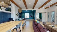 This thoughtfully designed single-floor space combines casual comfort with functional versatility across several distinct zones. Exposed wood beams span the white ceiling throughout, lending warmth and architectural character to the open layout. Light hardwood flooring unifies the space, complemented by an abundance of natural light streaming through large windows and an oversized garage-style glass door.

The lounge area serves as a central gathering point, anchored by a generous sectional sofa in deep burgundy accented with teal and yellow throw pillows. A shiplap-clad feature wall houses a linear fireplace with a concrete hearth and a wall-mounted television, flanked by arched built-in shelving with decorative displays. This area opens directly to an outdoor view, enhancing the sense of openness.

A kitchenette nearby features a striking blue herringbone tile backsplash, dark countertops, white cabinetry, and stainless steel appliances, topped with open floating shelves. A generous butcher-block communal table with wire-frame barstools provides a versatile workspace or casual dining area, illuminated by woven pendant lights.

The lounge transitions into a media and flex area defined by bold teal walls, a wall-mounted display, patterned area rug, and sliding barn doors that lead to a separate enclosed workspace, completing this dynamic, multi-purpose environment.