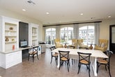 This single-floor community amenity space is thoughtfully designed to serve as both a welcoming social hub and a functional workspace. The layout is open and well-organized, seamlessly connecting several distinct areas under one roof.

At the front, oversized arched windows flood the lounge area with natural light and offer clear views of an outdoor pool and surrounding community. Elegant patterned drapery frames the windows, complementing a seating arrangement of plush, high-back wingback chairs in warm golden yellow tones paired with dark round accent tables and decorative lamps — creating an inviting atmosphere perfect for relaxed conversation.

Moving deeper into the space, the interior opens into a multipurpose community room featuring a bold dark shiplap accent wall that anchors the design. A built-in workstation with white cabinetry and open shelving provides a dedicated business center with a computer terminal and printer. Adjacent to this, a generous dining table with cross-back chairs offers a communal gathering area. A lounge nook with a banquette-style bench and a bold geometric area rug adds a cozy, residential feel.

Recessed lighting throughout ensures consistent, warm illumination. The neutral color palette of soft grays and whites, accented by yellow tones, creates a cohesive and modern aesthetic. The space is designed to accommodate both productivity and leisure for its community members.