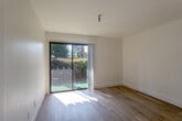 This efficiently designed one-bedroom, one-bathroom unit is laid out across a single floor, making smart use of every square foot. Light wood flooring runs throughout, creating a warm, cohesive feel from room to room. A hallway entry, accented by a wall-mounted sconce, leads into the main living area, which is bright and open thanks to a sliding glass door that frames views of a verdant, fenced outdoor space with mature palm trees and lush greenery.

The kitchen has been thoughtfully updated with crisp white shaker cabinets, white quartz countertops, a stainless undermount sink with a matte black faucet, a built-in induction cooktop, and an over-the-range microwave — all finished with sleek black hardware throughout.

The bedroom offers a comfortable footprint with a window overlooking outdoor greenery, providing ample natural light. A sliding barn-style closet door opens to a well-organized built-in closet with shelving and a hanging rod.

The bathroom features warm golden-yellow subway tile in the tub surround, beige floor tile, a white vanity with dark fixtures, a framed mirror, and stylish vanity lighting — adding a distinctive pop of color to the otherwise neutral palette. The overall ambiance is clean, bright, and move-in ready.