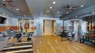 This fitness center offers a comprehensive workout environment designed to support a wide range of exercise routines. The space features warm hardwood flooring throughout, providing a clean and polished aesthetic that complements the functional layout. A dark, painted ceiling with multiple recessed lights and dual ceiling fans ensures the area remains well-lit and well-ventilated during workouts.

The cardio zone is outfitted with treadmills and an elliptical trainer, positioned along one side of the room for easy access. On the opposite side, a robust dumbbell rack with a full range of free weights and an adjustable bench provides ample options for strength training. A multi-station cable machine further expands the variety of exercises available.

Full-length mirrors line one wall, enhancing the sense of space and allowing users to monitor their form. The layout is open and thoughtfully organized, allowing for comfortable movement between equipment zones. Large glass-paneled doors and an adjacent room — visible through interior windows — suggest additional amenity spaces nearby, adding to the overall sense of a well-rounded facility. The overall ambiance is clean, modern, and inviting, reflecting a space purpose-built for fitness and wellness.
