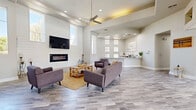Located at 1891 N Litchfield Rd in Goodyear, Arizona, this single-floor community space offers a bright, open, and welcoming environment designed with a clean, contemporary aesthetic. The layout flows seamlessly across one spacious level, blending multiple functional zones within an airy, well-connected interior.

The main living area is a standout feature, anchored by a striking shiplap accent wall that frames a sleek linear fireplace and wall-mounted television. Soaring ceilings with a ceiling fan overhead amplify the sense of openness, while tall vertical windows flood the room with natural light and offer views of the surrounding landscape. Light wood-look flooring runs throughout, complementing the neutral gray and white color palette.

An adjacent gathering area features a large communal table with bench seating, positioned near expansive windows that bring in ample daylight and connect the interior to the outdoors. A welcoming entry corridor showcases coastal-inspired artwork, lush greenery, and built-in planters, setting an inviting tone upon arrival.

Glass double doors lead to a fitness area, adding convenience and versatility to the overall space. Recessed lighting, tray ceiling details, and thoughtfully placed decorative accents—including coastal artwork and potted plants—round out the polished, resort-inspired atmosphere of this well-appointed community facility.