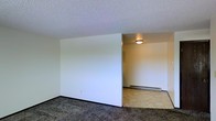 This single-floor space offers a straightforward and functional layout designed for comfortable everyday living. The main living area features dark, speckled carpet flooring that adds a layer of warmth underfoot, while a textured popcorn ceiling runs throughout, lending the space a classic, familiar character. Crisp white walls create a clean, neutral backdrop that allows for easy personalization and decorating.

An open passage connects the main living area to an adjoining room, which transitions to light-toned tile or vinyl flooring — a practical surface well-suited for high-traffic areas. This secondary space is equipped with a baseboard heating unit, ensuring efficient and consistent warmth throughout the colder months. A flush-mount ceiling light fixture provides steady, even illumination in this area.

The overall layout is open and unfussy, with a clear sightline between the two connected spaces that contributes to a sense of openness. The neutral color palette and simple finishes throughout make this a versatile space that can easily be adapted to a variety of uses and personal styles. The single-floor design ensures convenient, step-free access and navigation throughout the entire unit.