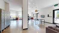 This single-floor amenity space in Walnut Creek, CA (94596) showcases a sleek, modern design characterized by polished concrete floors, crisp white walls, and high ceilings throughout. The space is divided into several distinct functional zones that flow seamlessly into one another.

The lounge area serves as a central gathering space, furnished with a large sectional sofa, a low-profile coffee table, and a woven area rug. A wall-mounted flat-screen display and a low media console anchor one wall, while roller shades filter natural light from expansive windows. A distinctive gold disc pendant light adds a warm, sculptural accent overhead.

Adjacent to the lounge is a well-equipped bike workshop and storage area. Polished floors reflect natural light from large windows, and the space accommodates bicycle racks, red tool chests, and a sturdy worktable on casters — providing a fully functional maintenance station for cyclists.

The entry and mailroom area features banks of individual metal mailboxes, stone-clad accent walls, and built-in bench seating with a perforated metal panel backdrop. A vibrant mural adds an artistic focal point near the entry corridor.

Throughout the space, recessed lighting and cassette-style HVAC units maintain a clean ceiling plane, reinforcing the contemporary, utilitarian aesthetic that defines this well-appointed communal facility.