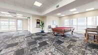 Located at 1000 Mount Pleasant in Toronto's midtown neighbourhood, this generously sized single-floor amenity space offers a versatile and well-appointed environment suited for gatherings, recreation, and socializing. The layout spans a substantial open floor plan, unified throughout by a bold patterned carpet that adds a distinctive design element to the neutral, warm-toned interiors.

The main open area is bright and airy, illuminated by a grid of recessed panel ceiling lights and flooded with natural light through expansive floor-to-ceiling windows that line the perimeter. These wide windows frame sweeping panoramic views of the Toronto skyline, serving as a striking visual backdrop throughout the space. Multiple seating vignettes are arranged to take advantage of these views, including lounge areas with a red accent armchair, upholstered sofa, and intimate bistro-style table seating.

A dedicated games area features a full-sized billiards table as a centrepiece, complemented by fitness accessories and additional seating. The space also includes a fully equipped kitchen with warm wood cabinetry, granite countertops, stainless steel appliances including a dishwasher and range, and a bay window offering city views. A wall-mounted television and ample circulation space make this amenity floor a comprehensive and functional hub for residents.