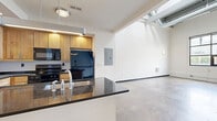 Located at 700 Everett St in Richmond, VA, this 2-bedroom, 2-bathroom loft spans three floors with a distinctly industrial aesthetic. The main level houses a combined dining, kitchen, and living area with polished concrete floors, exposed ductwork, and soaring ceilings that amplify the sense of space. The kitchen features a central island with dark granite countertops, a stainless steel sink, light wood cabinetry, and black appliances including a refrigerator, range, and built-in microwave. Large industrial-style windows on the main level flood the living area with natural light and offer views of the surrounding Richmond neighborhood. A striking steel staircase with wire mesh railings connects all three floors, serving as a focal point of the interior design. Exposed steel cross-bracing and structural beams reinforce the loft's industrial character throughout. The upper floors transition to warm hardwood flooring in the bedrooms, which feature high ceilings, clerestory windows, and dedicated closet space. Both bathrooms include light wood vanities with dark granite countertops and tile flooring. Additional conveniences include a dedicated laundry area and multiple closets. The space also includes a hallway connecting the upper-level rooms seamlessly.