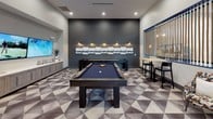This single-floor amenity lounge is thoughtfully designed to offer a premium recreational experience. The centerpiece of the space is a full-size pool table set atop a bold geometric-patterned carpet in shades of gray, white, and charcoal, which anchors the room and adds a striking visual element underfoot. A dramatic dark accent wall serves as a backdrop behind the pool table, adorned with a series of framed artwork and subtle wall-mounted lighting that creates an inviting, lounge-like atmosphere.

One of the most distinctive architectural features is the decorative rope-curtain partition, framed in navy blue, which visually connects the game room to an adjacent open lounge area. This design element adds texture and a nautical-inspired touch while maintaining an open, airy feel between zones.

A high-top table with bar stools is positioned near the rope divider, offering a casual seating area ideal for spectators or social gatherings. A wall-mounted flat-screen television nearby displays a golf simulation game, hinting at an integrated entertainment setup. The adjacent corridor transitions seamlessly to a hallway featuring wood-look flooring, gallery-style wall art, and additional seating, leading to a bright, open communal lounge beyond. Recessed lighting throughout ensures the space is well-lit and welcoming.