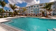 Located at 13370 Magnolia Oak Cir in North Fort Myers, Florida, this beautifully designed outdoor amenity space delivers a resort-inspired experience within a vibrant residential community. At the heart of the space is a large, sparkling swimming pool with a gradual entry, framed by wide paver pool decks and an array of lounge chairs perfect for soaking up the Florida sun. Tall palm trees are thoughtfully positioned throughout, offering natural shade and reinforcing the tropical ambiance.

Elegant cabanas with draped curtains and plush daybed seating are distributed around the pool perimeter, providing private retreats for relaxation. The surrounding landscape features vibrant tropical plantings, including bold red ti plants and lush green foliage that add vivid color contrast against the light-colored hardscape.

Beyond the pool area, manicured synthetic turf lawns serve as social gathering zones, complete with string lights overhead, cornhole boards, and casual seating arrangements ideal for outdoor entertaining. A dedicated gated garden area with paver walkways and colorful plantings adds a tranquil green retreat to the grounds.

The multi-story residential buildings, clad in coastal blue and gray hues with private balconies, frame the entire outdoor space, creating a cohesive and inviting community environment under the open Florida sky.