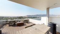 Located at 104 N Kenilworth Ave in Glendale, California, this rooftop terrace offers a well-appointed outdoor amenity space with sweeping panoramic views of the surrounding mountains and cityscape. The deck is finished with warm-toned composite wood planking throughout, creating a cohesive and durable surface that anchors the entire level.

The open-air section of the terrace is bordered by a sleek glass-panel railing system, maximizing unobstructed sightlines toward the Verdugo Mountains and the broader Glendale skyline. Multiple dining and seating arrangements are positioned across the deck to take full advantage of these views, accommodating a variety of gathering configurations.

A covered portion of the terrace provides a shaded retreat, complete with a wall-mounted flat-screen television and high-top bar seating — ideal for entertaining. Adjacent to this area, an outdoor kitchen is equipped with a stainless steel refrigerator, a built-in grill, and an extended granite-topped counter with a stainless steel sink, offering everything needed for al fresco dining and hosting.

Recessed ceiling lighting and security cameras are integrated throughout the covered zones, adding both functionality and a polished finish. Decorative planters with drought-tolerant greenery add organic touches to the space. The overall ambiance is modern and California-casual, combining clean architectural lines with an open-air setting that makes the most of the region's enviable climate and dramatic natural backdrop.