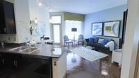 This thoughtfully designed single-floor space showcases a seamless open-concept layout that connects the kitchen, dining, and living areas into one cohesive, inviting environment. The kitchen features dark granite countertops, white cabinetry, and a built-in dishwasher, with pendant lighting in warm accent tones adding a refined touch overhead. A compact dining nook sits adjacent to the kitchen, accented by a round white table, decorative artwork, and a charming horse figurine that adds a curated, personal feel. The living area is bathed in natural light streaming through large windows dressed with yellow valances, creating a bright and airy atmosphere. Polished concrete floors run throughout, lending an industrial-chic character that complements the space's contemporary aesthetic. The living room is anchored by a deep charcoal sofa adorned with blue and green accent pillows, paired with classic patterned armchairs and a glass coffee table atop a patterned area rug. Framed abstract artwork lines the walls, contributing to the space's artistic sensibility. A floor lamp and side table complete the sitting area, making it a comfortable and visually cohesive retreat. Overall, the space balances modern finishes with classic furnishings for an elegant yet livable ambiance.