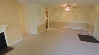 This single-floor residence offers a straightforward and functional layout designed for comfortable everyday living. The main living area is generously proportioned, featuring wall-to-wall carpet in a neutral beige tone that contributes to a warm and understated ambiance throughout. Walls are painted in a soft, light hue with a two-tone treatment — a slightly deeper tan shade along the lower portion separated by a subtle chair rail — adding a modest architectural detail to the otherwise clean interior. A ceiling fan with an integrated light fixture is centrally mounted, providing both air circulation and ambient lighting to the open space. A structural column or partial wall divides the living area and leads the eye toward a connected kitchen, visible through a pass-through opening. The kitchen features warm wood cabinetry and appears to include standard appliances, offering a functional cooking and meal preparation space. White baseboards frame the floors throughout, providing a crisp contrast to the carpeted surfaces. The overall design is practical and move-in ready, with a cohesive neutral palette that provides a versatile backdrop for a variety of furnishing styles.