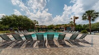 This outdoor amenity area centers around a large, well-maintained swimming pool set within a generously sized paver-stone deck designed for both relaxation and socializing. Rows of slatted lounge chairs are arranged along the pool's perimeter, offering ample space for sunbathing beneath the open Florida sky. Tall palm trees and mature surrounding greenery provide a tropical ambiance throughout the space.

Adjacent to the pool, a single-story clubhouse building serves as the focal point of the complex. The structure features a striking exterior combining bold orange panel siding, exposed brick columns, and large floor-to-ceiling windows that allow natural light to flow freely. A covered outdoor patio area with high-top tables and chairs extends from the building, creating a shaded lounge space ideal for gathering. A roll-up garage-style door on one side suggests flexible indoor-outdoor functionality, while a secure glass entry door provides access to the interior.

A covered gazebo pavilion is visible in the background near the pool's far end, offering additional shaded seating. The entire area is enclosed by a fence, ensuring privacy and safety. The combination of the paver deck, tropical landscaping, and well-appointed amenity structures gives this outdoor space a resort-like character suited for an active, community-oriented lifestyle.