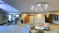This single-floor space is thoughtfully designed to offer a warm and inviting communal environment. The lounge area immediately draws attention with its distinctive tray ceiling, accentuated by a circular recessed design and multiple recessed spotlights that cast a soft, even glow throughout the room. The flooring features light-toned, wood-style planks that contribute to the airy, modern feel of the space.

A standout feature is the stone-clad fireplace wall, which anchors one end of the room with a rustic yet refined aesthetic. Mounted above the fireplace is a flat-screen television, making this corner a natural focal point for both relaxation and socializing. Adjacent to the fireplace, a reception or service desk area is partially visible, suggesting this space serves a communal or hospitality-oriented function.

The seating arrangement is central to the room's layout, with patterned upholstered armchairs grouped around a round white coffee table, creating an intimate conversation area. The bold circular print on the chairs adds a contemporary design element that contrasts pleasantly with the neutral wall tones.

Warm sconce lighting on the walls complements the overhead recessed fixtures, enhancing the overall ambiance. A wood-trimmed doorway with elevator call panels nearby indicates convenient access to other areas, rounding out this well-appointed and functional communal lounge.
