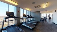 This elevated fitness center is thoughtfully designed to support a full range of workout activities within a modern, well-appointed space. The facility is organized into distinct functional zones, each tailored to specific training needs. The cardio area lines a bank of floor-to-ceiling windows, positioning a row of Life Fitness treadmills and elliptical machines to take full advantage of expansive panoramic city views, flooding the space with natural light throughout the day. Dark rubber flooring throughout the cardio and strength zones provides both durability and a sleek, professional aesthetic.

The strength training zone is generously equipped with a comprehensive selection of resistance machines, free weights, barbells, weight racks, and adjustable benches. Large mirrors line the walls, visually expanding the space and allowing users to monitor their form. A centrally located accessible restroom serves the facility conveniently.

A separate studio room features light wood-look flooring, floor-to-ceiling windows admitting abundant natural light, and storage for stability balls and accessories, making it well-suited for stretching, yoga, or group fitness. Glass partitions and doors connect the studio to the main floor, maintaining an open, airy feel while providing acoustic separation. The overall ambiance is clean, modern, and purposeful.
