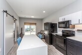 This single-floor unit showcases a contemporary design with a clean, neutral palette and light wood-look flooring that runs throughout the space, creating a cohesive and modern aesthetic. The kitchen is a standout feature, equipped with black appliances including a refrigerator, electric range, over-the-range microwave, and dishwasher. Dark lower cabinetry contrasts sharply with white upper cabinets and light countertops, while under-cabinet lighting adds a polished touch. A freestanding island with open shelving below provides additional counter space and casual seating for two.

The living area is compact yet functional, featuring a sliding glass door that invites natural light and offers a view of a landscaped outdoor area. The bedroom is simply appointed, accommodating a platform bed with built-in storage drawers and a window with a roller shade for privacy.

A notably versatile bathroom and laundry corridor runs alongside the bedroom, housing a stacked washer and dryer unit, a toilet, and a vanity with a white countertop and dark wood cabinetry. Recessed lighting and a clean, minimalist finish carry through each area of the unit, contributing to an overall atmosphere that is functional, modern, and well-organized.