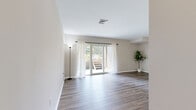 This single-floor space offers a clean, modern aesthetic defined by neutral tones, wood-look flooring, and an open layout that promotes a sense of spaciousness throughout. The living area is a standout feature, highlighted by sliding glass doors dressed with sheer white curtains that invite abundant natural light and provide direct access to an outdoor deck, offering views of surrounding greenery.

The kitchen is well-appointed with white shaker-style cabinetry, granite countertops, a white subway tile backsplash, and stainless steel appliances including a gas range and dishwasher. A corner sink adds a functional design element, while a peninsula with bar seating creates a natural transition into the open living and dining area.

The bedrooms feature light wood-look flooring and large windows that allow natural light to filter in, creating bright, airy spaces. Each room includes closet access and maintains the same neutral color palette found throughout the unit.

The bathroom features classic diamond-patterned tile flooring and a shower-tub combination. A stacked washer and dryer unit is conveniently located nearby, adding in-unit laundry functionality. Overall, the space presents a cohesive, well-maintained interior with practical amenities and a welcoming ambiance.