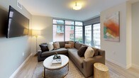 This contemporary single-floor residence showcases a thoughtfully designed layout that balances comfort and modern aesthetics throughout. The living area is a standout space, featuring expansive floor-to-ceiling windows that wrap around the corner, flooding the room with natural light and offering open views of the surrounding urban streetscape. Light hardwood flooring runs throughout, lending warmth and continuity to each room.

The kitchen is equipped with stainless steel appliances, including a side-by-side refrigerator, range, and microwave, all complemented by sleek white high-gloss cabinetry, dark wood lower cabinets, granite countertops, and under-cabinet lighting, creating a clean, high-end culinary space.

The bedrooms are generously proportioned, each featuring large windows that bring in ample daylight and views of the surrounding neighborhood. Light wood flooring, neutral wall tones, and built-in closet storage contribute to a calm, well-organized environment.

The bathrooms reflect a consistent modern design language, featuring large-format gray textured wall tiles accented by vertical strips of smaller mosaic tiles. The primary bathroom includes a walk-in shower with a frameless glass panel and a linear drain, while the secondary bathroom offers a full soaking tub and shower combination. Both spaces are finished with clean white fixtures and chrome hardware.
