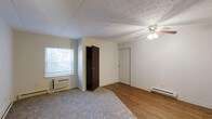 This single-floor, 2-bedroom, 1-bathroom unit offers a straightforward and functional layout designed for comfortable everyday living. The living area features a combination of hardwood flooring and carpeting, creating distinct zones within the open space. A ceiling fan with a built-in light fixture provides both ventilation and ambient lighting, while a window-mounted air conditioning unit ensures climate control. A baseboard heater along the wall offers an additional heating source during cooler months. The textured ceiling adds a subtle design detail to the room. The bedroom area is well-appointed with two closets — one standard closet and one open storage closet — providing generous storage options. A window dressed with horizontal blinds allows natural light to filter in while maintaining privacy, with views of surrounding greenery visible from inside. The neutral wall tones throughout the unit create a clean, versatile backdrop that adapts easily to various interior styles. Overall, the space presents a practical and well-equipped living environment with essential amenities integrated thoughtfully into its compact, single-level footprint.