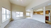 This 2-floor space offers a thoughtfully designed open-concept main living area that immediately impresses with its vaulted ceilings, creating a generous sense of vertical space and airiness. The living room is finished with wall-to-wall carpeting in a neutral gray tone, providing a comfortable and cohesive foundation throughout the space. Smooth, beige-toned walls keep the aesthetic clean and versatile.

Two windows along the front wall allow natural light to filter into the room, brightening the interior throughout the day. A ceiling fan is positioned overhead in the living area, adding both functionality and a casual, practical element to the design.

The layout transitions naturally into a secondary area with standard ceiling height, where a pass-through counter connects to the kitchen. The kitchen features warm oak cabinetry and overhead fluorescent lighting, offering a functional and straightforward cooking space. A door visible in the background likely leads to additional rooms or storage.

The combination of open living space, vaulted ceilings, and a practical kitchen layout makes this a functional and comfortable multi-floor residence.
