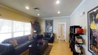 This single-floor residence offers a thoughtfully designed interior that balances comfort and style. The living room serves as the heart of the space, showcasing warm hardwood-style flooring that runs throughout and creates a cohesive, inviting atmosphere. Recessed lighting is installed across the ceiling, providing even, well-distributed illumination, while crown molding along the ceiling line adds a refined architectural detail. The room features rich, dark leather seating arranged around a substantial dark wood coffee table, anchored by a patterned area rug that adds texture and visual interest. A large decorative art print serves as a bold focal point on one wall, lending personality to the space. Natural light filters in through a window dressed with layered curtains, complementing the warm neutral tones of the walls. A tall bookcase along one side of the room offers display and storage space, while a decorative indoor plant adds a touch of greenery. A mirrored door leads to an adjoining area, enhancing the sense of depth and openness. The overall ambiance is warm, curated, and functional, reflecting a well-appointed single-level living space.
