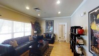 This single-floor residence offers a thoughtfully designed interior that balances comfort and style. The living room serves as the heart of the space, showcasing warm hardwood-style flooring that runs throughout and creates a cohesive, inviting atmosphere. Recessed lighting is installed across the ceiling, providing even, well-distributed illumination, while crown molding along the ceiling line adds a refined architectural detail. The room features rich, dark leather seating arranged around a substantial dark wood coffee table, anchored by a patterned area rug that adds texture and visual interest. A large decorative art print serves as a bold focal point on one wall, lending personality to the space. Natural light filters in through a window dressed with layered curtains, complementing the warm neutral tones of the walls. A tall bookcase along one side of the room offers display and storage space, while a decorative indoor plant adds a touch of greenery. A mirrored door leads to an adjoining area, enhancing the sense of depth and openness. The overall ambiance is warm, curated, and functional, reflecting a well-appointed single-level living space.