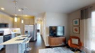 This well-appointed single-floor residence offers a thoughtfully designed open-concept layout that seamlessly connects the living and kitchen areas. The living space features warm neutral walls, dark hardwood flooring, and a cozy seating area anchored by a mid-century modern media console and a bold accent chair in rich terracotta orange, adding a pop of color to the refined palette. Framed wall art and a soft area rug further enhance the inviting ambiance.

The kitchen is outfitted with light wood cabinetry that extends to the ceiling, providing generous storage. A stainless steel French door refrigerator with an ice and water dispenser serves as a focal point, complemented by a peninsula-style breakfast bar with white countertops and modern bar seating — ideal for casual dining. Pendant lighting adds a warm, functional glow over the counter space.

A hallway visible from the main living area leads to additional rooms, hinting at a well-organized floor plan. Recessed lighting throughout keeps the space bright and modern, while large windows allow natural light to filter in. The overall aesthetic blends contemporary design with functional comfort, creating a space that feels both stylish and livable.