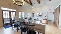 This single-story clubhouse blends rustic charm with modern comfort through a thoughtfully designed open-concept layout. The space is anchored by soaring vaulted ceilings adorned with prominent exposed wood beams and finished with white shiplap, creating a warm yet airy atmosphere. A wrought-iron wagon wheel chandelier serves as a striking focal point above the dining area, reinforcing the rustic aesthetic throughout.

The dining area features two substantial reclaimed wood farmhouse tables paired with classic black Windsor chairs, providing generous seating for gatherings. Large multi-pane windows line the walls, inviting abundant natural light and offering views of the surrounding exterior. Light-toned hardwood flooring runs seamlessly throughout, further enhancing the sense of openness.

Adjacent to the dining space, a comfortable lounge area is outfitted with upholstered seating and a wall-mounted flat-screen television, making it well-suited for casual socializing or community events. Recessed lighting and adjustable wall-mounted fixtures supplement the natural light, ensuring the space is well-lit at any hour. The overall ambiance is welcoming, functional, and designed to encourage community connection.