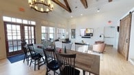 This single-story clubhouse blends rustic charm with modern comfort through a thoughtfully designed open-concept layout. The space is anchored by soaring vaulted ceilings adorned with prominent exposed wood beams and finished with white shiplap, creating a warm yet airy atmosphere. A wrought-iron wagon wheel chandelier serves as a striking focal point above the dining area, reinforcing the rustic aesthetic throughout.

The dining area features two substantial reclaimed wood farmhouse tables paired with classic black Windsor chairs, providing generous seating for gatherings. Large multi-pane windows line the walls, inviting abundant natural light and offering views of the surrounding exterior. Light-toned hardwood flooring runs seamlessly throughout, further enhancing the sense of openness.

Adjacent to the dining space, a comfortable lounge area is outfitted with upholstered seating and a wall-mounted flat-screen television, making it well-suited for casual socializing or community events. Recessed lighting and adjustable wall-mounted fixtures supplement the natural light, ensuring the space is well-lit at any hour. The overall ambiance is welcoming, functional, and designed to encourage community connection.