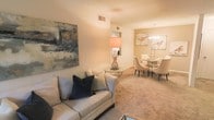 This single-floor residence offers a thoughtfully designed layout that seamlessly blends comfort and elegance. The open-concept living and dining area serves as the heart of the home, creating a welcoming and cohesive environment ideal for both relaxing and entertaining. The living area is anchored by plush wall-to-wall carpeting in warm neutral tones, providing a cozy foundation for the stylishly furnished space. A large, bold abstract artwork adorns one wall, adding a striking visual element and a touch of contemporary flair. A glass-topped side table and a softly glowing table lamp contribute to the refined yet approachable ambiance. The transition into the dining area is fluid and natural, where a round dining table surrounded by upholstered chairs sits beneath an elegant chandelier, lending a sense of formal sophistication to the space. Soft, warm lighting throughout enhances the inviting atmosphere. A mirrored accent in the corner adds depth and dimension to the interior. The neutral color palette, composed of soft beiges and whites, ties the entire space together, creating a harmonious and timeless aesthetic that is both functional and visually appealing.