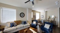 This single-floor space presents an open, flowing layout that connects the living and dining areas in a cohesive design. The living room is anchored by a comfortable beige sofa adorned with decorative throw pillows and a warm knit blanket, complemented by a pair of navy blue accent chairs with nailhead trim detailing. A dark wood coffee table sits atop a light area rug, grounding the seating arrangement. Dark hardwood floors extend throughout, providing a rich contrast to the soft, neutral wall tones. A ceiling fan with integrated lighting fixture serves as a functional focal point overhead, while recessed lighting adds additional illumination throughout the space. Multiple windows dressed with horizontal blinds invite natural light while maintaining privacy. A decorative wall clock adds character to the living area. The adjoining dining space is visible through a subtle architectural column divide, featuring additional seating and wall décor, including a framed American flag motif. The overall ambiance is warm and inviting, blending traditional and transitional design elements for a comfortable, lived-in aesthetic.