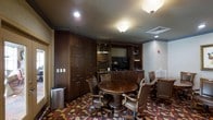 This distinguished meeting room exudes a classic, executive ambiance through its carefully curated design elements and rich finishes. The centerpiece of the space is a substantial round wooden table surrounded by high-backed leather executive chairs, creating an intimate yet professional setting well-suited for meetings, discussions, or private gatherings. Floor-to-ceiling dark wood cabinetry lines the walls, offering generous storage and lending the room a warm, refined character. Built-in shelving units flank a recessed media area equipped with a flat-screen display, making the space equally functional for presentations. A wet bar or kitchenette counter runs along the back wall, providing added convenience for extended sessions. The room is illuminated by flush-mount ceiling fixtures that cast a warm, focused glow throughout. A boldly patterned carpet in deep reds, golds, and dark tones grounds the space and adds visual depth, complementing the rich wood tones of the cabinetry and furnishings. Crown molding details along the upper walls add an architectural finishing touch. The single-floor layout is efficiently organized, balancing comfort and functionality in a compact, elegantly appointed environment.