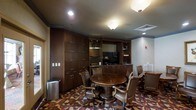 This distinguished meeting room exudes a classic, executive ambiance through its carefully curated design elements and rich finishes. The centerpiece of the space is a substantial round wooden table surrounded by high-backed leather executive chairs, creating an intimate yet professional setting well-suited for meetings, discussions, or private gatherings. Floor-to-ceiling dark wood cabinetry lines the walls, offering generous storage and lending the room a warm, refined character. Built-in shelving units flank a recessed media area equipped with a flat-screen display, making the space equally functional for presentations. A wet bar or kitchenette counter runs along the back wall, providing added convenience for extended sessions. The room is illuminated by flush-mount ceiling fixtures that cast a warm, focused glow throughout. A boldly patterned carpet in deep reds, golds, and dark tones grounds the space and adds visual depth, complementing the rich wood tones of the cabinetry and furnishings. Crown molding details along the upper walls add an architectural finishing touch. The single-floor layout is efficiently organized, balancing comfort and functionality in a compact, elegantly appointed environment.