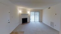 This single-floor residence offers a clean, understated aesthetic with a welcoming and functional layout. The living area serves as the heart of the space, centered around a classic wood-burning fireplace framed by a crisp white mantel, adding a timeless focal point to the room. Soft, neutral-toned carpeting extends throughout, creating a warm and comfortable underfoot experience. Wall-mounted sconce lighting on either side of the fireplace provides a gentle, ambient glow that complements the natural light streaming in through large sliding glass doors fitted with vertical blinds. These doors not only flood the space with daylight but also offer direct access to an outdoor area, seamlessly connecting the interior to the exterior. The walls are finished in a clean, light neutral tone that enhances the sense of openness and brightness throughout. The overall ambiance is calm and versatile, providing a blank canvas well-suited for a variety of interior styles and furnishing arrangements. The single-floor layout ensures easy accessibility and a cohesive flow throughout the space, making it both practical and comfortable for everyday living.