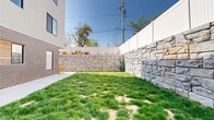 This thoughtfully designed outdoor space serves as a shared exterior area for a contemporary multi-story building. The grounds are divided into two distinct zones: a landscaped green area and a functional hardscape section. The lawn area is enclosed by striking natural stone retaining walls, which vary in texture and tone — ranging from lighter cut limestone to darker, rougher-faced granite blocks — creating a visually dynamic boundary. A white vinyl privacy fence tops the perimeter, adding an additional layer of seclusion under open blue skies. The hardscape zone features a broad, clean concrete patio that provides ample room for movement and gathering. A warm-toned wooden pergola anchors this area, offering a shaded focal point alongside a row of dark, multi-unit cluster mailboxes set against the stone wall backdrop. Young trees have been planted throughout, adding greenery and a sense of growth to the space. The adjacent building showcases a modern architectural aesthetic, combining dark brick cladding at the base with light stucco, wood accent panels, and large contemporary windows on the upper floors. Individual garage doors with frosted glass panels line the ground level, contributing to the building's clean, urban character. Together, these elements create a practical yet attractive outdoor environment.