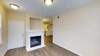 This single-floor residence offers a thoughtfully arranged layout that balances comfort and functionality. The living area serves as the heart of the space, anchored by a classic white-mantled fireplace that adds warmth and a welcoming focal point to the room. A built-in niche above the fireplace provides an architectural detail that adds character to the otherwise clean, neutral walls finished in soft warm tones. Hardwood-style flooring runs throughout, lending a sense of continuity and warmth across the open floor plan. Natural sightlines connect the living area to the kitchen, which is visible in the background and features light-toned cabinetry, hinting at a bright and functional cooking space. The layout promotes an open, airy flow between living and culinary areas, making the space well-suited for both everyday living and casual entertaining. Flush-mount ceiling lighting provides even illumination throughout, while HVAC vents integrated into the ceiling and walls ensure year-round comfort. White baseboards and trim provide crisp contrast against the flooring and walls, contributing to a polished, well-maintained appearance throughout the residence.