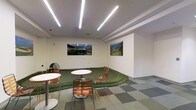 This single-floor space is designed as a recreational and social area centered around an indoor putting green. The green is the defining feature of the room, with a curved, organically shaped boundary that sets it apart from the surrounding tiled flooring. The contrast between the artificial turf and the checkered tile creates a visually dynamic layout that anchors the space.

Overhead, linear LED lighting strips are arranged in a diagonal pattern across the white ceiling, providing bright, even illumination throughout the room. The clean white walls serve as a neutral backdrop for two framed photographic prints depicting lush golf course landscapes, reinforcing the golf theme and adding a sense of depth and color to the otherwise minimal decor.

Seating is provided by several lightweight metal-framed chairs with slatted wooden seats, paired with small round pedestal tables. These are positioned both on the putting green and on the tiled surround, encouraging casual conversation and relaxed interaction.

The overall ambiance is clean, modern, and purposeful — balancing a functional recreational element with a comfortable lounge atmosphere, making it well-suited for both active use and informal social engagement.
