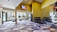 Located at 483 Scott Blvd in Castle Rock, Colorado, this single-floor fitness center is designed to support a full range of workout routines in a well-appointed, energizing environment. The main exercise area is defined by soaring vaulted ceilings and bold golden-yellow accent walls that give the space a vibrant, motivating atmosphere. A wall-mounted flat-screen television is positioned high on the feature wall, visible from across the room. The geometric-patterned carpet in shades of grey and white adds a dynamic visual element to the floor throughout the facility. Cardio equipment, including treadmills, ellipticals, and stationary bikes, is arranged along a bank of large windows that flood the space with natural light and offer views of the surrounding community. A dedicated free weights area includes a multi-tier dumbbell rack and medicine ball storage tower, accompanied by floor mirrors that enhance the sense of openness. An adjoining hallway connects to restrooms and additional amenity spaces, with colorful artwork decorating the walls and a water fountain conveniently available. A door from the cardio area opens directly to an outdoor patio, providing easy access to fresh air. The overall design balances functionality with an inviting aesthetic, creating a comfortable setting for daily fitness activity.
