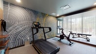 Located at 30 W First Ave in Columbus, Ohio, this well-appointed fitness center offers a thoughtfully designed single-floor workout space that balances functionality with modern aesthetics. The room's most striking feature is a bold, large-scale mural accent wall rendered in charcoal gray and gold, depicting concentric ring patterns reminiscent of a tree's cross-section — a visually dynamic backdrop that energizes the space.

The cardio zone is equipped with a treadmill and Peloton-style cycling bikes, positioned on protective floor mats. Adjacent to this area, a dedicated strength and flexibility zone features hardwood flooring, a wall-mounted ballet barre, full-length framed mirrors, and an adjustable workout bench — ideal for stretching, barre workouts, or weight training.

Floor-to-ceiling sheer curtains line an expansive window wall, allowing generous natural light to filter in while offering views of an outdoor patio area. Recessed ceiling lighting throughout ensures the space remains bright and functional at any time of day. The entry area includes a wooden storage unit with hooks, a waste bin, and posted fitness guidelines, providing practical organization. Neutral gray carpeting in the cardio zone and warm wood-tone flooring in the training alcove create a clear visual distinction between workout areas, contributing to an overall clean, contemporary atmosphere.