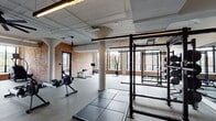 This fitness facility occupies a single floor within a building of clear industrial heritage, where original exposed brick walls, structural concrete columns, and open ceiling systems with visible ductwork establish a distinctive architectural character. Large, black-framed windows are positioned throughout, flooding the space with natural light and offering street-level views of the surrounding neighborhood.

The main workout floor is expansive and open-plan, housing a comprehensive selection of cardio equipment including Xebex rowing machines, treadmills, ellipticals, and stationary bikes. A dedicated strength training zone features power racks with weight plates, cable machines, a Smith machine, and free weight storage. Rubber flooring and interlocking mats define the lifting areas, while large ceiling fans and linear LED lighting maintain comfort throughout.

A separate studio space features hardwood floors, floor-to-ceiling mirrors, a wall-mounted flat-screen display, and glass sliding doors. It is stocked with stability balls, medicine balls, resistance bands, and yoga mats. An additional alcove provides space for functional training with step platforms and a dumbbell rack. The entry corridor features polished concrete floors, pendant lighting, and exposed brick, providing a cohesive transition into the facility.