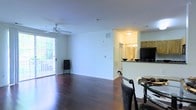 This single-floor residence offers a thoughtfully designed open-concept layout that seamlessly connects the main living area to the kitchen and dining space. The living room is grounded by rich, dark hardwood flooring that runs throughout, lending warmth and elegance to the space. A ceiling fan overhead provides comfort, while a large sliding glass door floods the room with natural light and opens onto an outdoor deck, offering pleasant views of surrounding greenery.

The open layout creates a natural flow into the kitchen area, which is defined by a raised breakfast bar with a dark countertop that serves as both a functional workspace and a casual dining spot. The kitchen is equipped with classic wooden cabinetry in a warm honey tone, providing ample storage, and features a full-size refrigerator. Recessed and flush-mount lighting ensures the kitchen remains bright and inviting.

Clean white walls throughout the living and kitchen areas create a neutral, versatile backdrop that complements the darker flooring and countertops. The overall ambiance of the space is open, airy, and move-in ready, making it an ideal setting for both everyday living and casual entertaining.